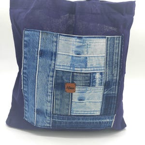 Peut inclure: Un sac fourre-tout en toile bleu foncé avec une grande poche en patchwork de denim sur le devant. La poche est faite de différentes nuances de denim bleu et porte une étiquette en cuir marron avec le mot "Mimao" écrit dessus.