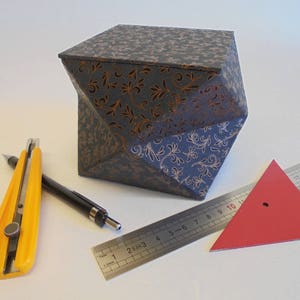 Könnte beinhalten: Eine dunkelblaue Box mit goldenem Blumenmuster, ein gelbes Cuttermesser, ein schwarzer Bleistift, ein silbernes Lineal und ein rotes Dreieck.