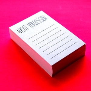 Peut inclure: Une pile de papier à lettres blanc avec des lignes horizontales et le texte allemand "NICHT VERGESSEN" imprimé en noir. Le papier à lettres est sur un fond rose vif.
