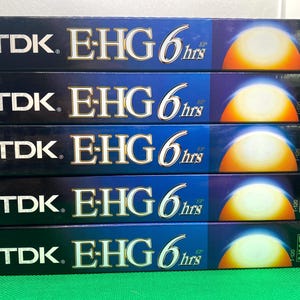 May include: A stack of five TDK E-HG VHS video cassette tapes. The tapes are black with blue and gold accents, featuring the TDK logo and "E-HG 6 hrs" text. Each tape has a sunset-like graphic and VHS label.