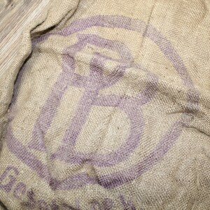 May include: A brown burlap sack with a purple circular logo. The logo has a stylized letter "G" inside.