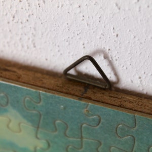 K&ouml;nnte beinhalten: Nahaufnahme eines Holzrahmens mit einem Metall-Aufh&auml;nger. Der Rahmen h&auml;lt ein Puzzle mit einer blauen und gr&uuml;nen Himmelsszene.