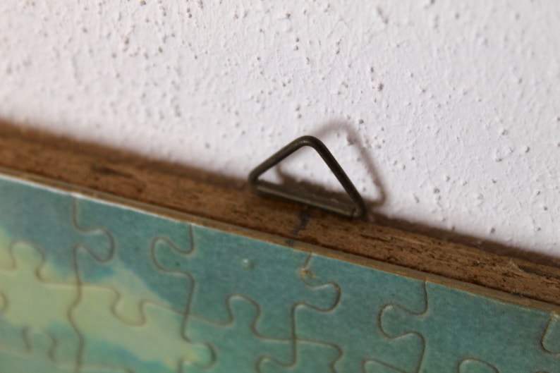 K&ouml;nnte beinhalten: Nahaufnahme eines Holzrahmens mit einem Metall-Aufh&auml;nger. Der Rahmen h&auml;lt ein Puzzle mit einer blauen und gr&uuml;nen Himmelsszene.