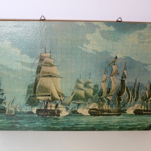 K&ouml;nnte beinhalten: Ein gerahmtes Vintage-Puzzle, das eine maritime Szene mit mehreren Segelschiffen auf einem blauen und gr&uuml;nen Meer darstellt. Die Schiffe haben wei&szlig;e Segel und befinden sich in einer Schlachtformation. Der Himmel ist hellblau mit wei&szlig;en Wolken.