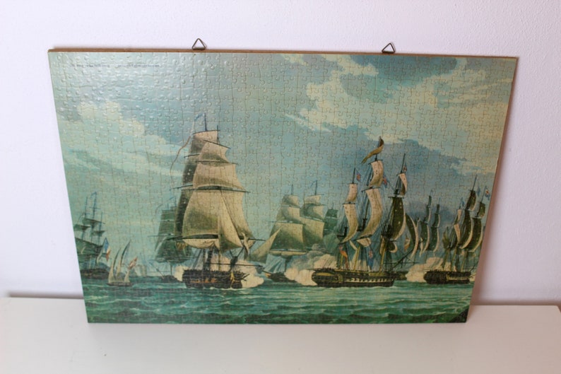 K&ouml;nnte beinhalten: Ein gerahmtes Vintage-Puzzle, das eine maritime Szene mit mehreren Segelschiffen auf einem blauen und gr&uuml;nen Meer darstellt. Die Schiffe haben wei&szlig;e Segel und befinden sich in einer Schlachtformation. Der Himmel ist hellblau mit wei&szlig;en Wolken.