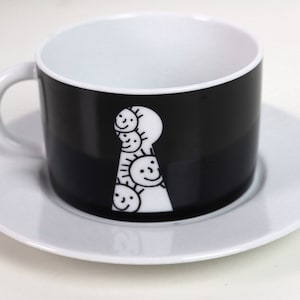 May include: A black and white teacup and saucer set with a keyhole design featuring a stack of smiling faces.