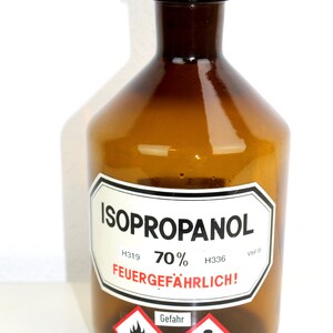 Puede incluir: Una botella de vidrio marr&oacute;n con una tapa negra, etiquetada "ISOPROPANOL 70% H319 H336 VbF B FEUERGEF&Auml;HRLICH!" con un s&iacute;mbolo de llama roja y blanca y un signo de exclamaci&oacute;n.
