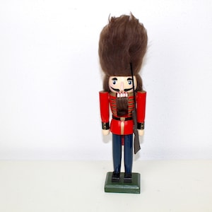 Könnte beinhalten: Ein traditionelles Nussknacker-Spielzeug aus Holz mit einer rot-goldenen Uniform, einem braunen Pelzhut und einem schwarzen Schnurrbart. Der Nussknacker hält ein Gewehr in seiner rechten Hand.