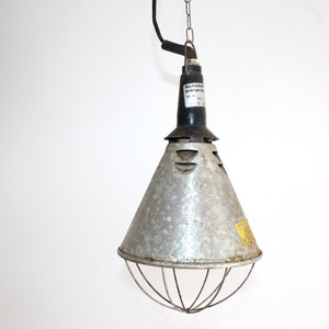 May include: A vintage industrial-style hanging lamp with a metal conical shade and a protective wire cage. The lamp has a black top section and is suspended by a chain. A label is visible on the lamp.