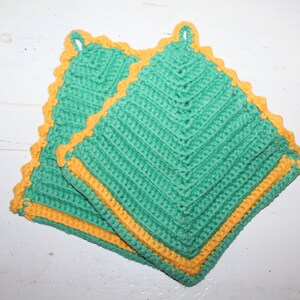 Könnte beinhalten: Zwei handgefertigte Topflappen aus Häkelarbeit. Die Topflappen sind quadratisch, mit einer türkisfarbenen Mitte und einem gelben Rand. Jeder Topflappen hat eine Schlaufe zum Aufhängen. Der Häkelstich erzeugt eine strukturierte, gemusterte Oberfläche.