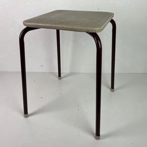 Könnte beinhalten: Ein kleiner, quadratischer Hocker mit einer hellgrauen Sitzfläche und dunkelbraunen Metallbeinen. Die Beine sind leicht gebogen und haben silberfarbene Kappen an der Unterseite. Der Hocker ist ein einfaches, funktionales Möbelstück.