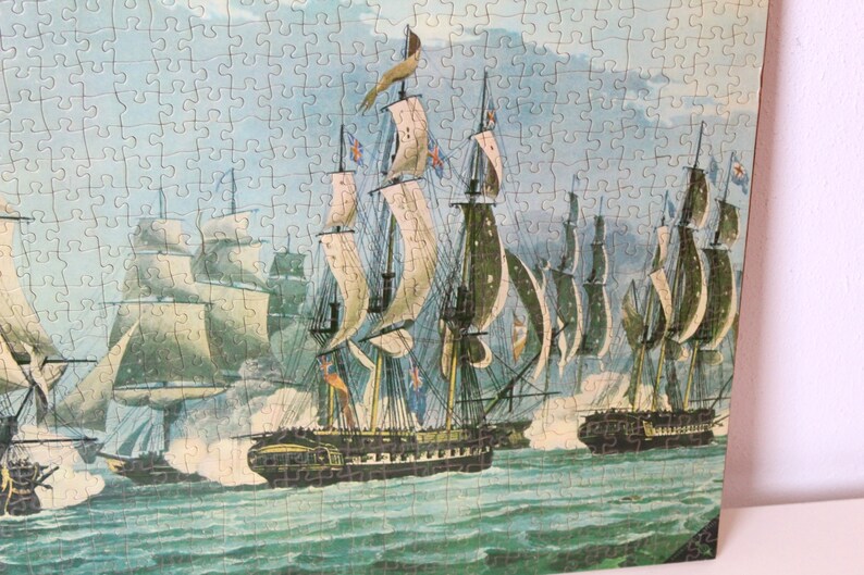 K&ouml;nnte beinhalten: Ein Vintage-Puzzle, das eine Seeschlacht mit mehreren Segelschiffen im Ozean darstellt. Die Schiffe haben wei&szlig;e Segel und feuern Kanonen aufeinander ab. Der Himmel ist blau und bew&ouml;lkt.