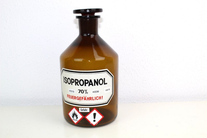 Puede incluir: Una botella de vidrio marr&oacute;n con una etiqueta blanca que dice "ISOPROPANOL 70% FEUERGEF&Auml;HRLICH!" con un s&iacute;mbolo de advertencia rojo y blanco. La botella tiene una tapa de pl&aacute;stico negro.
