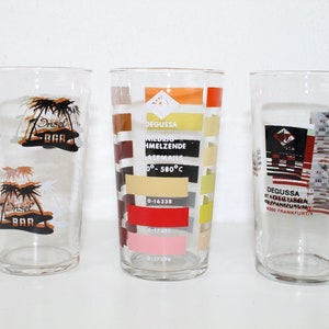 Könnte beinhalten: Drei klare Glas-Trinkgläser mit unterschiedlichen Designs. Das erste Glas hat ein Palmenmotiv mit dem Text "Insel Bar". Das zweite Glas hat ein farbenfrohes geometrisches Design mit dem Text "Degussa Schmelzende Emaille 0-580°C 0-16338". Das dritte Glas hat ein schwarz-rotes geometrisches Design mit dem Text "Degussa KDS Glasba Panseum 6000 Frankfurt/M".