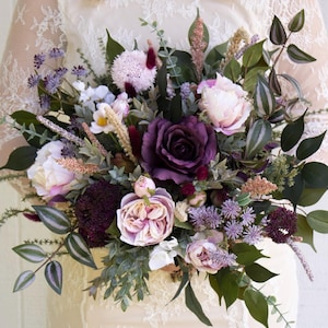 May include: A large, lush bouquet featuring a variety of flowers in shades of purple, pink, and white, with green foliage. The bouquet includes roses, peonies, and other floral elements. The bouquet is held in front of a white background.