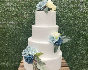 May include: A four-tier white cake dummy decorated with blue and white artificial flowers. The cake is displayed on a wooden crate.