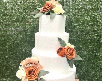 May include: A four-tier white cake decorated with orange, peach, and cream-colored artificial flowers. The cake is on a wooden crate.