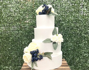 May include: A four-tier white wedding cake decorated with white and blue flowers. The cake is sitting on a wooden crate.