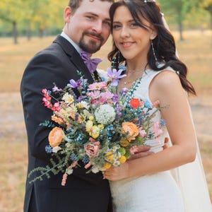 K&ouml;nnte beinhalten: Ein Paar posiert f&uuml;r ein Foto, die Frau in einem wei&szlig;en Hochzeitskleid und Schleier, der Mann in einem schwarzen Anzug mit einer lila Fliege. Die Frau h&auml;lt einen bunten Blumenstrau&szlig; mit rosa, orangefarbenen und blauen Bl&uuml;ten.