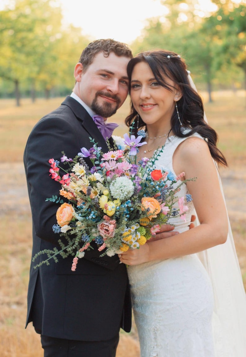 K&ouml;nnte beinhalten: Ein Paar posiert f&uuml;r ein Foto, die Frau in einem wei&szlig;en Hochzeitskleid und Schleier, der Mann in einem schwarzen Anzug mit einer lila Fliege. Die Frau h&auml;lt einen bunten Blumenstrau&szlig; mit rosa, orangefarbenen und blauen Bl&uuml;ten.