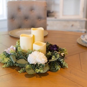 May include: A centerpiece with three ivory pillar candles surrounded by a green wreath with white and purple flowers. The centerpiece is on a wooden table.