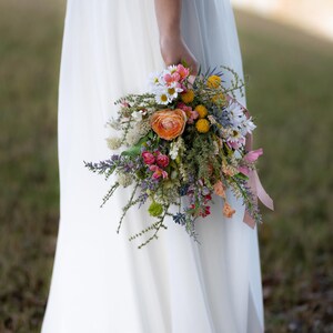 K&ouml;nnte beinhalten: Ein lebendiger Strau&szlig; Wildblumen, darunter orangefarbene Rosen, G&auml;nsebl&uuml;mchen und Lavendel, gehalten vor einem wei&szlig;en Kleid. Die Anordnung ist mit einem rosa Band gebunden und schafft eine nat&uuml;rliche und farbenfrohe Pr&auml;sentation, ideal f&uuml;r eine Hochzeit.