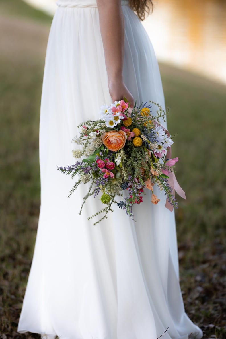 K&ouml;nnte beinhalten: Ein lebendiger Strau&szlig; Wildblumen, darunter orangefarbene Rosen, G&auml;nsebl&uuml;mchen und Lavendel, gehalten vor einem wei&szlig;en Kleid. Die Anordnung ist mit einem rosa Band gebunden und schafft eine nat&uuml;rliche und farbenfrohe Pr&auml;sentation, ideal f&uuml;r eine Hochzeit.