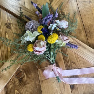 K&ouml;nnte beinhalten: Ein Strau&szlig; k&uuml;nstlicher Blumen mit gelben, lila, wei&szlig;en und rosa Bl&uuml;ten, gebunden mit einem hellrosa Band. Die Anordnung umfasst Lavendel und G&auml;nsebl&uuml;mchen. Der Strau&szlig; liegt auf einer Holzoberfl&auml;che.
