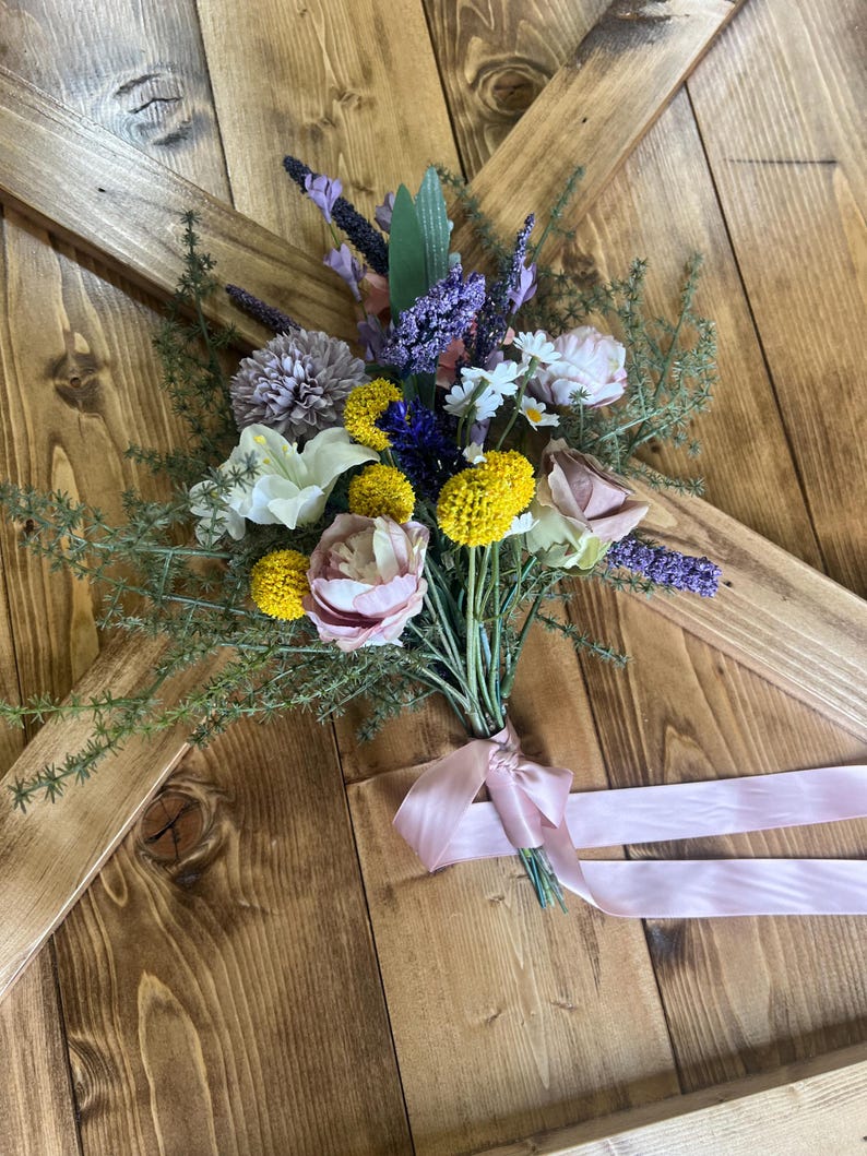K&ouml;nnte beinhalten: Ein Strau&szlig; k&uuml;nstlicher Blumen mit gelben, lila, wei&szlig;en und rosa Bl&uuml;ten, gebunden mit einem hellrosa Band. Die Anordnung umfasst Lavendel und G&auml;nsebl&uuml;mchen. Der Strau&szlig; liegt auf einer Holzoberfl&auml;che.