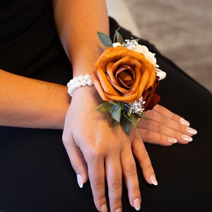 Peut inclure: Une corsage de poignet avec une grande rose orange, des fleurs blanches et des feuilles vertes. La corsage est attach&eacute;e avec un bracelet de perles.