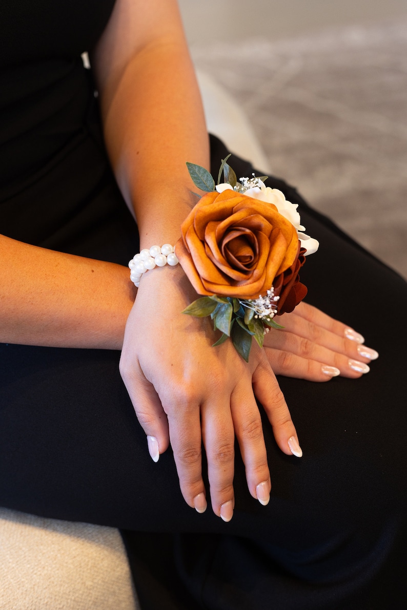 Peut inclure: Une corsage de poignet avec une grande rose orange, des fleurs blanches et des feuilles vertes. La corsage est attach&eacute;e avec un bracelet de perles.