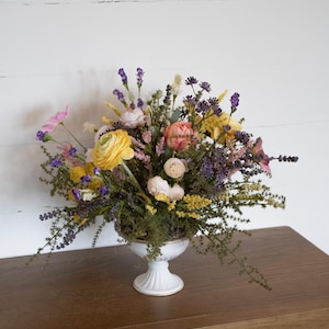 K&ouml;nnte beinhalten: Ein lebendiger Blumenstrau&szlig; in einer wei&szlig;en Sockelvase. Der Strau&szlig; zeigt gelbe, rosa und lila Blumen mit Gr&uuml;n. Die Vase steht auf einer braunen Holzoberfl&auml;che vor einer wei&szlig;en Wand.