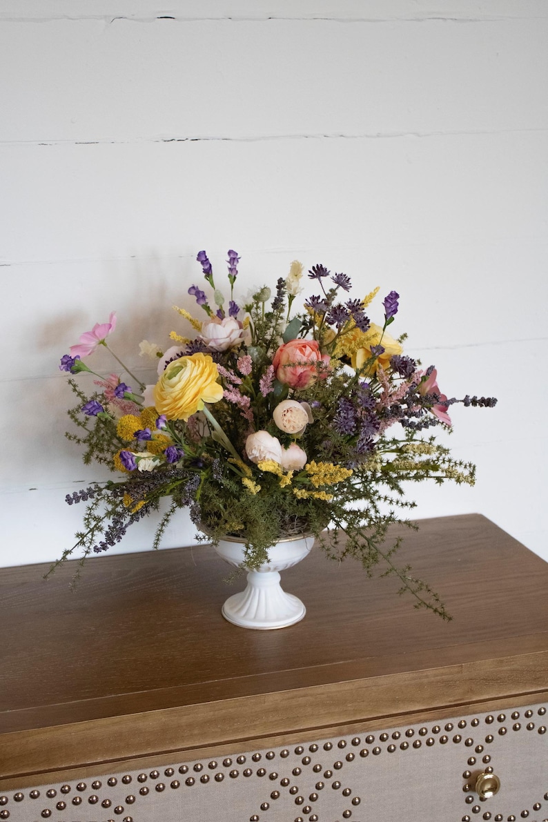 K&ouml;nnte beinhalten: Ein lebendiger Blumenstrau&szlig; in einer wei&szlig;en Sockelvase. Der Strau&szlig; zeigt gelbe, rosa und lila Blumen mit Gr&uuml;n. Die Vase steht auf einer braunen Holzoberfl&auml;che vor einer wei&szlig;en Wand.