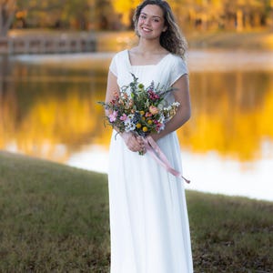 K&ouml;nnte beinhalten: Eine Frau in einem wei&szlig;en Kleid h&auml;lt einen bunten Blumenstrau&szlig; mit einem rosa Band. Das Kleid hat einen quadratischen Ausschnitt und kurze &Auml;rmel. Der Hintergrund zeigt einen See und B&auml;ume mit goldgelbem Laub.