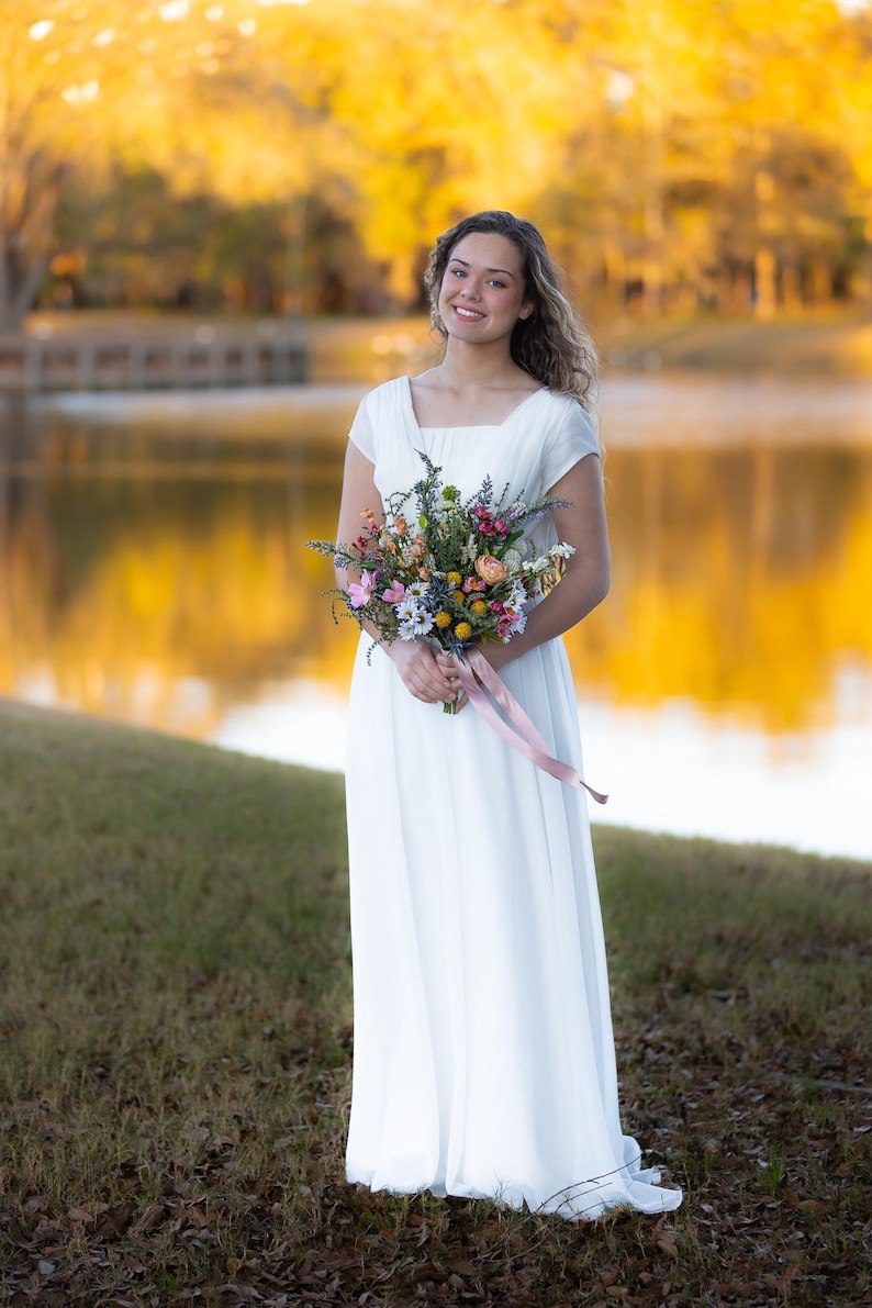 K&ouml;nnte beinhalten: Eine Frau in einem wei&szlig;en Kleid h&auml;lt einen bunten Blumenstrau&szlig; mit einem rosa Band. Das Kleid hat einen quadratischen Ausschnitt und kurze &Auml;rmel. Der Hintergrund zeigt einen See und B&auml;ume mit goldgelbem Laub.