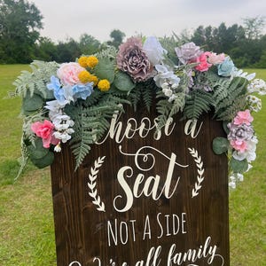 K&ouml;nnte beinhalten: Ein dunkelbraunes Holzschild mit wei&szlig;er Schrift, auf dem steht: "W&auml;hle einen Platz, keine Seite, wir sind alle Familie". Das Schild ist mit einem Blumengesteck aus rosa, blauen, gelben und wei&szlig;en Blumen und gr&uuml;nem Laub geschm&uuml;ckt.
