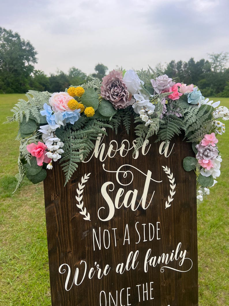 K&ouml;nnte beinhalten: Ein dunkelbraunes Holzschild mit wei&szlig;er Schrift, auf dem steht: "W&auml;hle einen Platz, keine Seite, wir sind alle Familie". Das Schild ist mit einem Blumengesteck aus rosa, blauen, gelben und wei&szlig;en Blumen und gr&uuml;nem Laub geschm&uuml;ckt.