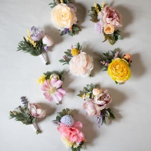 K&ouml;nnte beinhalten: Verschiedene florale Corsagen und Boutonnieren in Pfirsich-, Rosa-, Gelb- und Lavendelt&ouml;nen. Jede Anordnung weist verschiedene Blumensorten und Gr&uuml;n auf. Text auf dem Bild: Jede Corsage und Boutonniere variiert.