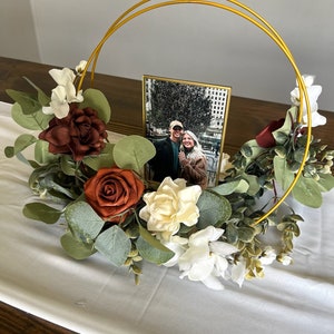 May include: A gold metal hoop with a picture frame inside. The frame holds a photo of a couple. The hoop is surrounded by a floral arrangement with white, red, and green flowers.