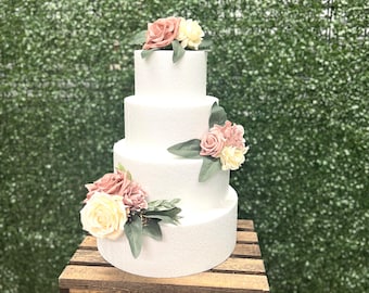 May include: A four-tier white cake decorated with pink and cream flowers. The cake is on a wooden crate in front of a green wall.