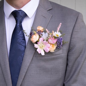 K&ouml;nnte beinhalten: Ein grauer Anzug mit einem wei&szlig;en Hemd und einer marineblauen Paisley-Krawatte. Eine florale Ansteckblume mit rosa, pfirsichfarbenen, wei&szlig;en und lila Blumen ist am Revers des Anzugs befestigt. Die Blumen setzen einen Farbakzent.