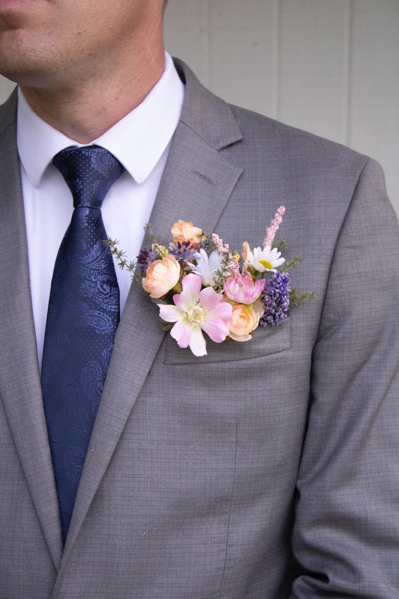 K&ouml;nnte beinhalten: Ein grauer Anzug mit einem wei&szlig;en Hemd und einer marineblauen Paisley-Krawatte. Eine florale Ansteckblume mit rosa, pfirsichfarbenen, wei&szlig;en und lila Blumen ist am Revers des Anzugs befestigt. Die Blumen setzen einen Farbakzent.