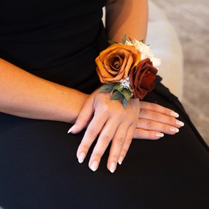 Peut inclure: Une corsage de poignet avec trois roses dans des tons de brun, orange et rouge. Les roses sont dispos&eacute;es en grappe avec de la verdure et de petites fleurs blanches.