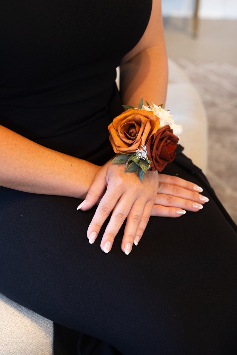 Peut inclure: Une corsage de poignet avec trois roses dans des tons de brun, orange et rouge. Les roses sont dispos&eacute;es en grappe avec de la verdure et de petites fleurs blanches.