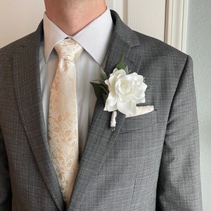 May include: A grey suit jacket with a white shirt and a gold patterned tie. A white flower with green leaves is pinned to the lapel.