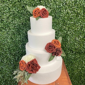 May include: A four-tiered white cake decorated with faux rust-colored roses, orange roses, and white flowers. Green leaves accent the floral arrangements. The cake sits on a wooden stand against a green hedge backdrop.