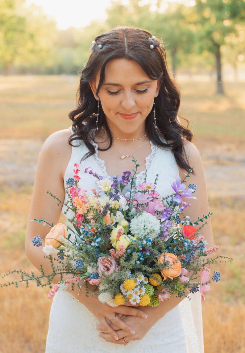 K&ouml;nnte beinhalten: Ein farbenfroher Blumenstrau&szlig; mit einer Vielzahl von Farben, darunter Rosa, Orange, Gelb und Blau. Der Strau&szlig; wird von einer Person in einem wei&szlig;en Kleid gehalten. Der Hintergrund ist eine verschwommene Szene im Freien.