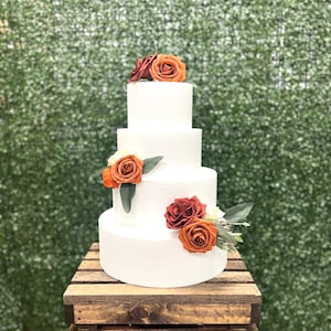 May include: A four-tier white wedding cake decorated with orange and burgundy roses and greenery. The cake is sitting on a rustic wooden crate.