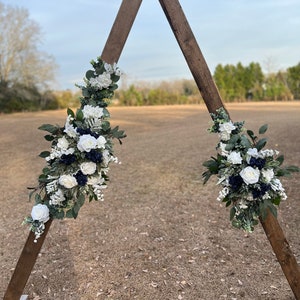 May include: A wooden triangular archway decorated with white and blue floral arrangements. The archway is standing in a field of brown grass.