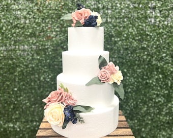 May include: A four-tier white wedding cake decorated with pink and cream roses and blue flowers. The cake is on a wooden crate.